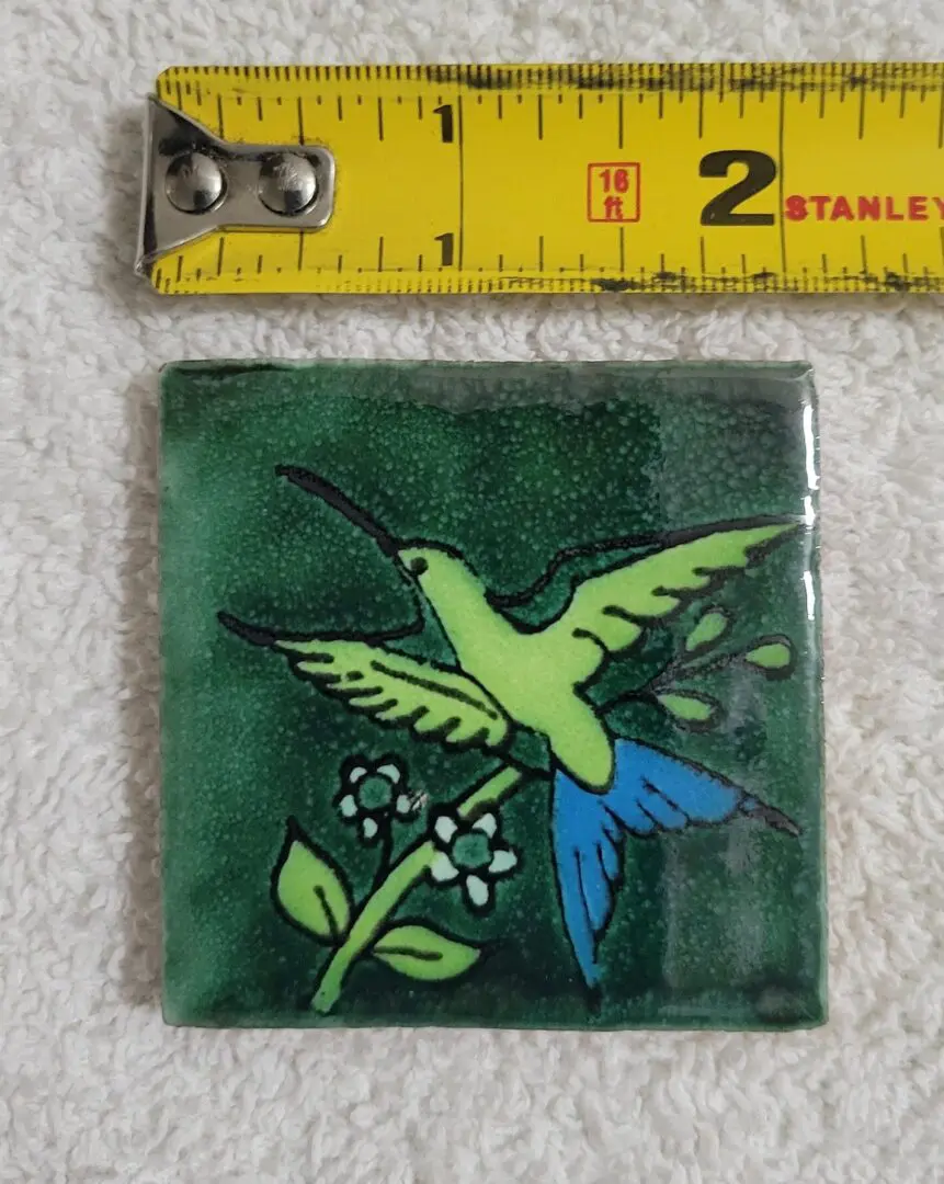 Green tile with a hummingbird and flower design next to a measuring tape.