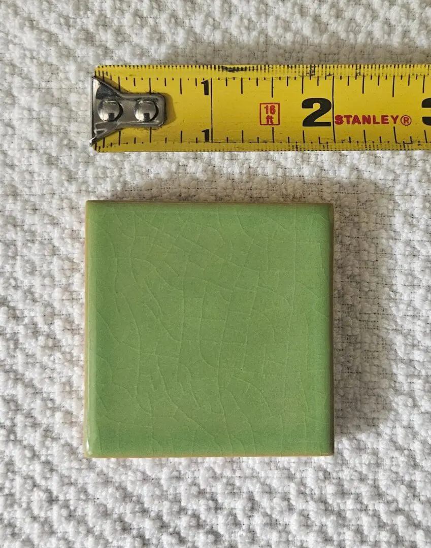 A green square tile next to a measuring tape showing about 2 inches.