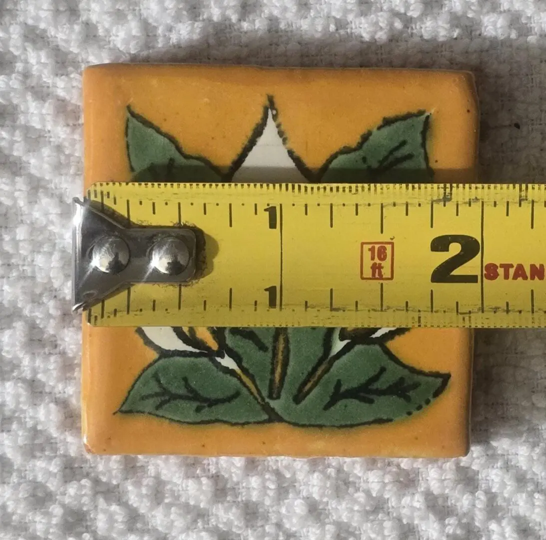 A small square ceramic tile with a green leaf design and measuring tape showing its size.