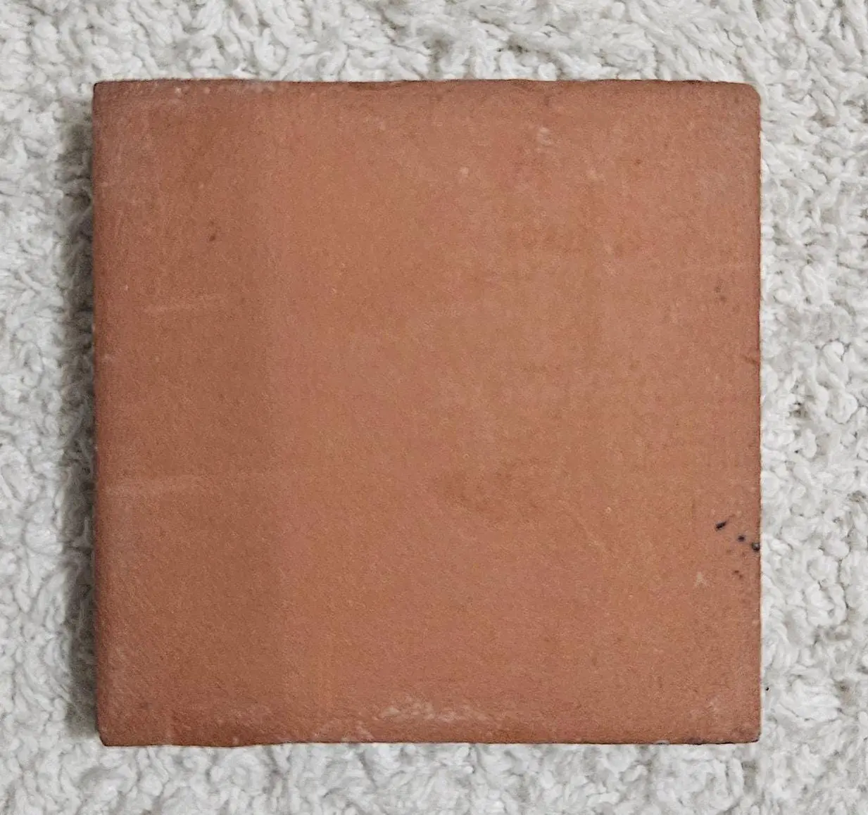 A single square terracotta tile on a speckled surface.