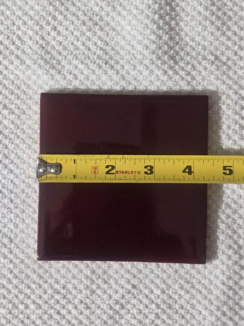 A dark red square tile with a measuring tape showing about 3.5 inches.