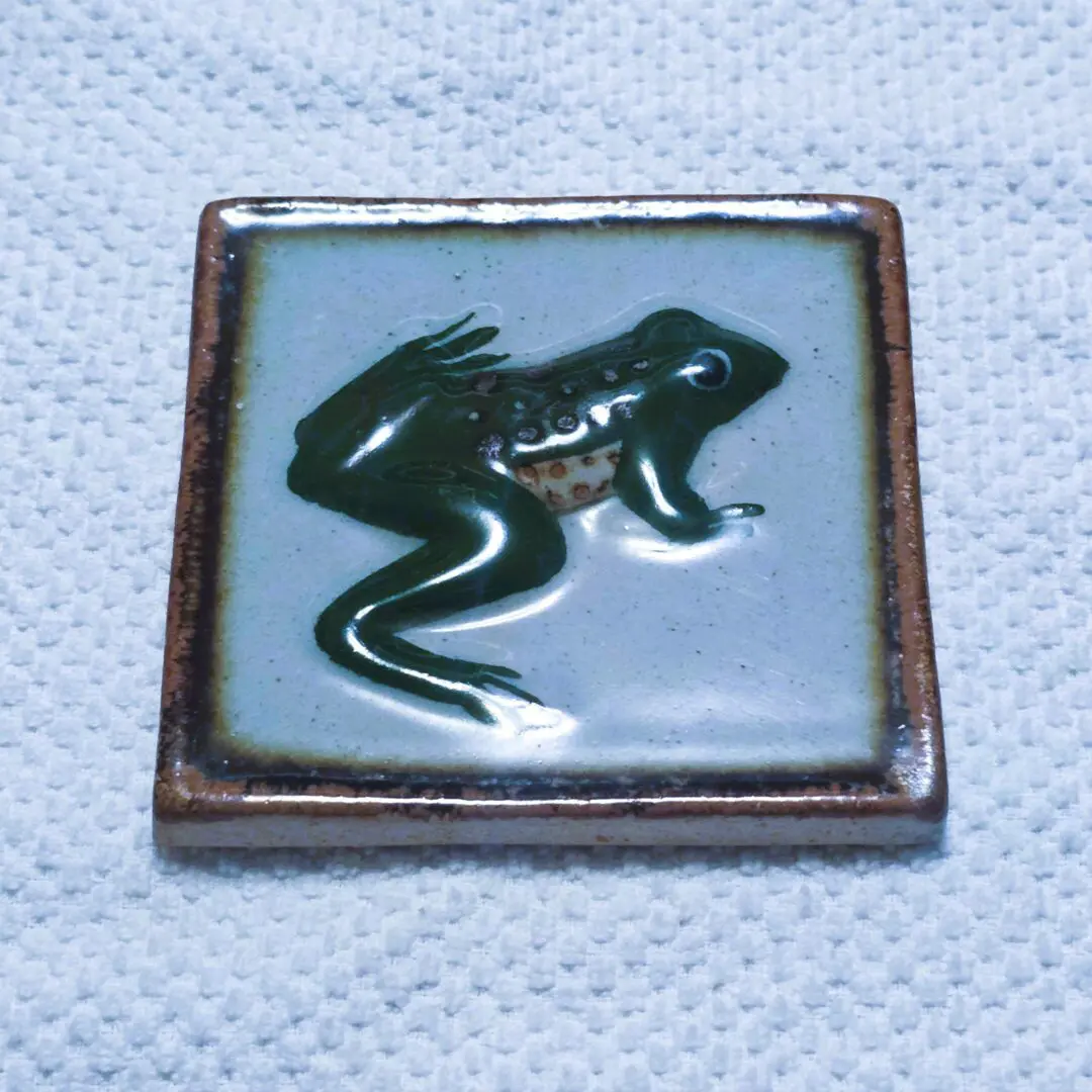 A ceramic tile with a green frog design on a white background.