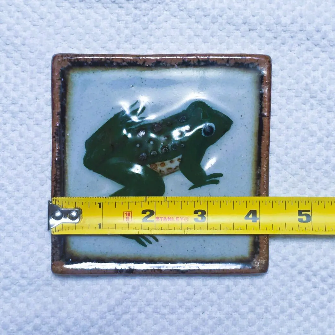 Square tile with a green frog design and measuring tape.