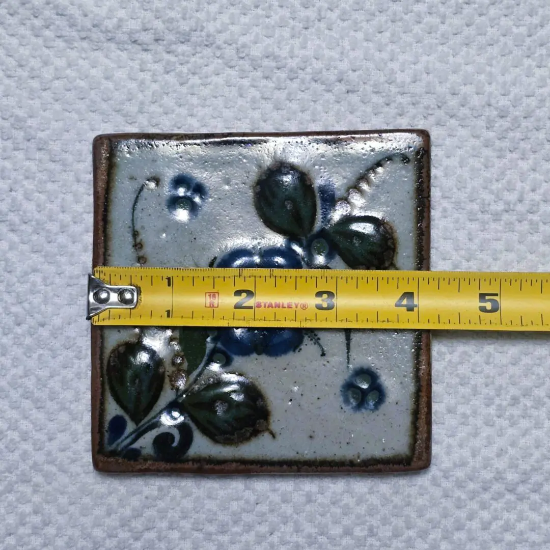 A small square ceramic tile with a floral design and a measuring tape across it.