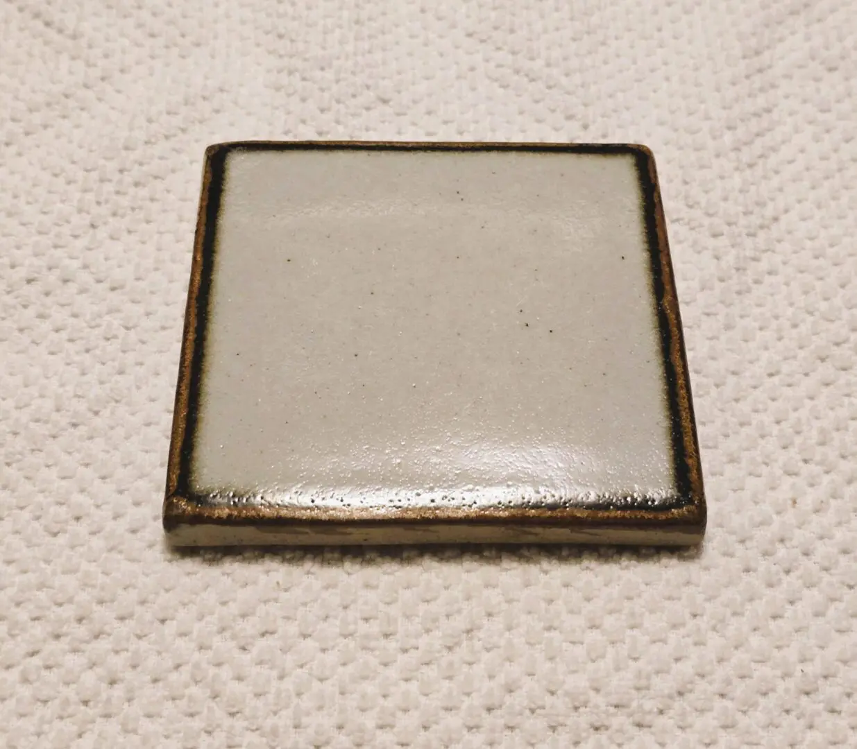 A small square ceramic tile with a plain beige surface and brown edges.