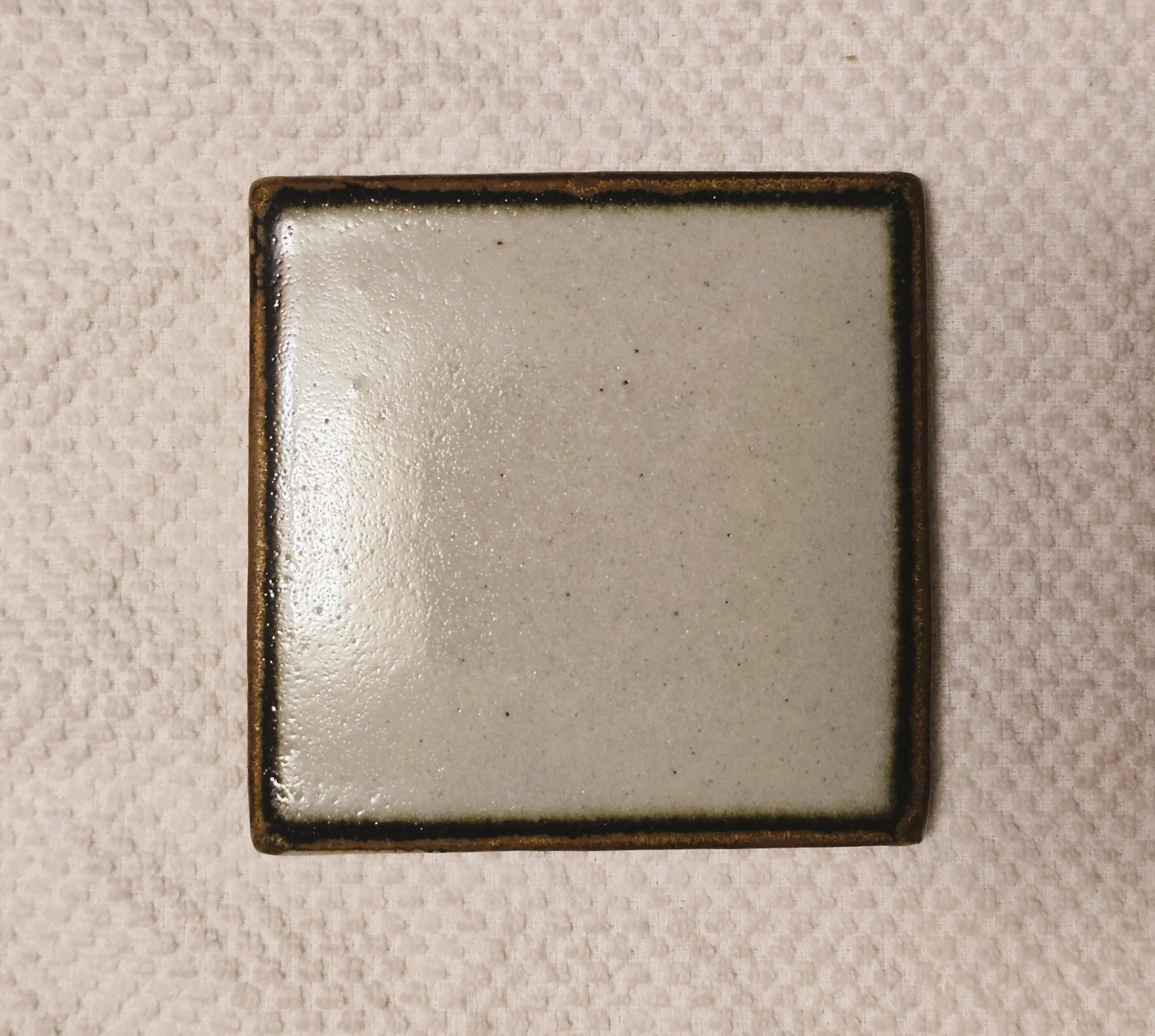 A square ceramic tile with a dark border on a textured surface.