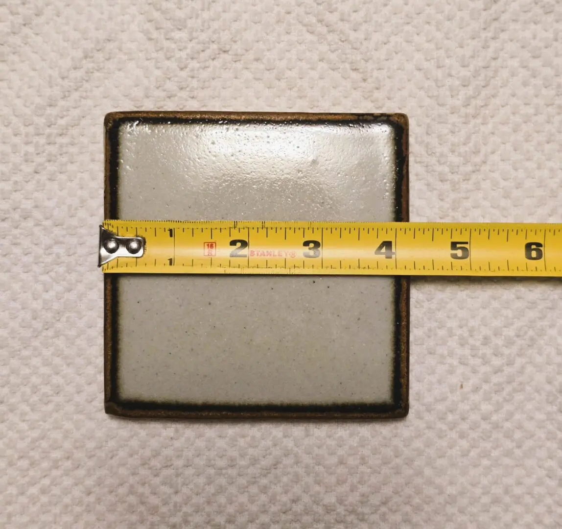A square ceramic tile with a measuring tape showing its width.